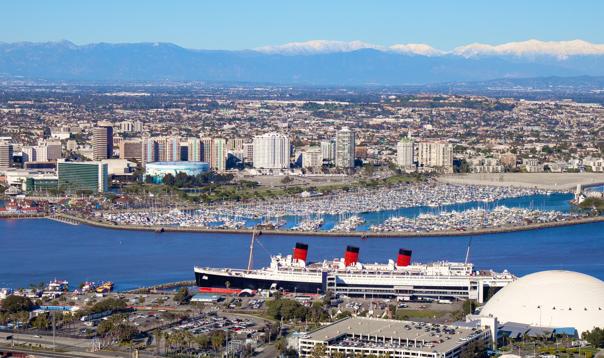 Aerial view of the Long Beach waterfront with the Queen Mary, marina, downtown skyline, and snow-capped mountains